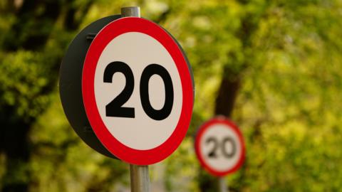 A generic image of a red and white 20mph speed limit sign on a blurry tree background. A second sign, slightly blurred, can be seen behind the first, more prominent sign.