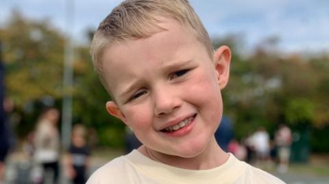 Ozzy, a little boy with blond hair, stands smiling with his head titled to the side. 