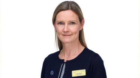 A woman with long fair hair looks straight at the camera in a professional head shot. She is wearing an NHS name badge on a blue top