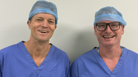 Glanso UK Directors, Dr Matthew Molyneux and Dr Neil Rasburn look at the camera and smile. They are wearing blue NHS uniforms, with hair nets.