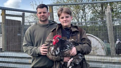 Tom, a man with short brown hair and brown top, with his son, also called Tom, who has brown hair, a brown jacket and blue top, holding a cockerel. There is a wooden and mesh cage behind them in an allotment and some trees.