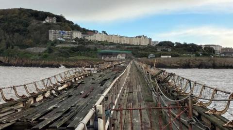 A view from Birnbeck Pier - a disused and damaged Victorian pier, which is missing many of its wooden flooring planks and is crumbling, looking back towards Weston-super-Mare. Houses and woodland can be seen in the distance.