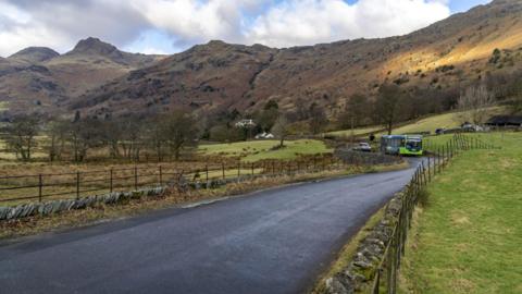 A bus pictured in the Lake District. The bus is coming round a corner and the fells are visible in the background.