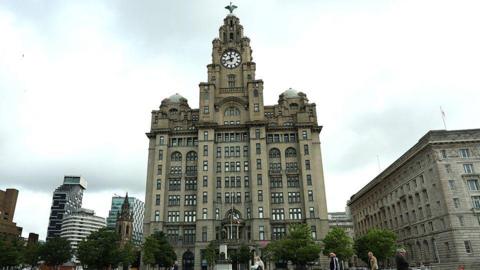People walking past the Royal Liver Building by the docks in Liverpool on a dry day.