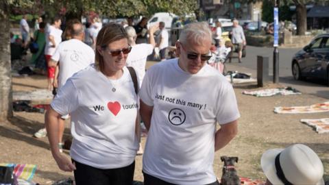 A man and woman in white t-shirts looking down. One top says: 'we love tourists', the other days: 'but not this many'