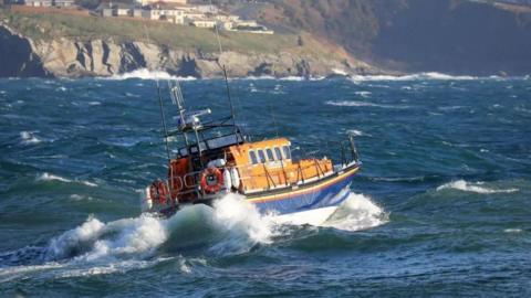 Douglas lifeboat sailing in a choppy sea. The boat is in the RNLI blue, orange and white colours and is leading to the left. Buildings can be seen on a headland in the background.