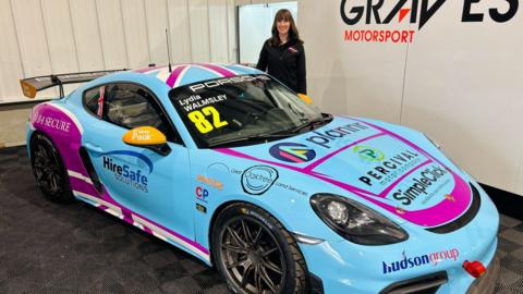 Lydia Walmsley dressed in all black stands in a car show room with her blue Porsche.