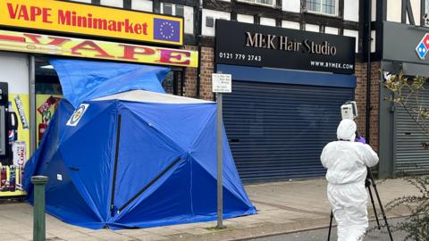 A row of shops. A vape minimarket shop with a yellow and red sign has a blue forensics tent attached to its front entrance. There is a forensics worker in a white protective suit holding a camera on a tripod