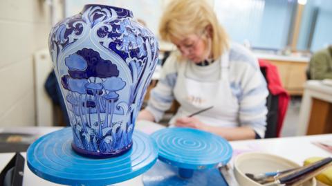 A newly-made blue vase with a pattern of trees on it has been set on a table. A woman with blonde hair and glasses can be seen sitting down and painting in the background.