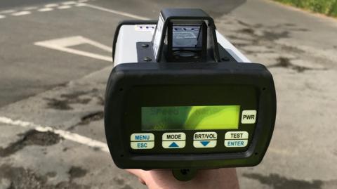 A close-up of a person's hand holding a traffic speed enforcement device. The road surface in the background is uneven and contains potholes.