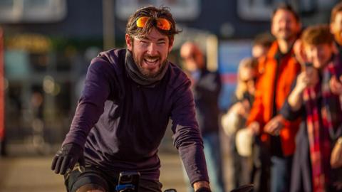 Nathan Hurley is riding his bike at Cardiff Bay in front of a crowd. He has dark hair and a beard and has a big grin