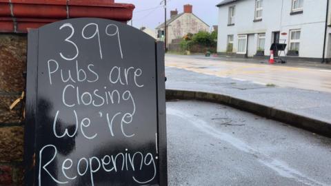 A pub sign next to a busy main road - It reads "399 pubs are closing , we're reopening !" 
