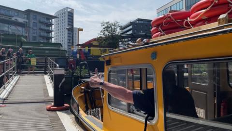 A bright yellow water taxi in Leeds. A man has his arm out of the window of the boat giving a thumbs up sign towards a group of onlookers standing at the entrance to a jetty. Office and apartment tower blocks can be scene in the background against a cloudy sky.