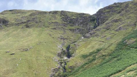 A narrow waterfall carved into the side of a steep and rocky fell. The slope is covered in grass with various rock outcrops. The waterfall curves down the side of it. The sky is blue with thin clouds.