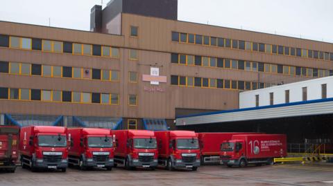 A row of red postal lorries are lined up outside Cardiff sorting office. The building has brown cladding on and horizontal rows of windows with the Royal Mail insignia on the front. 
