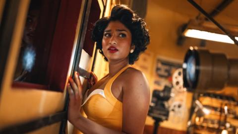 Varada Sethu in a promo image from Doctor Who where she is dressed in a yellow top and is wearing red lipstick.