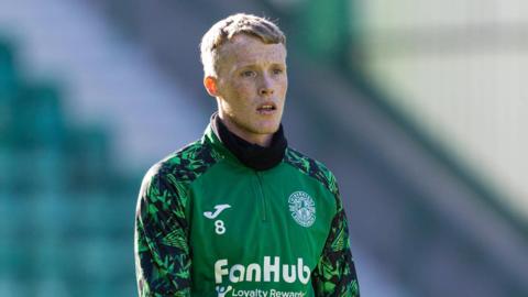 Jake Doyle-Hayes wearing Hibs training gear. He is wearing a green top with the Hibs badge in the top right corner. He is wearing long green and black sleeves and black gloves. He has fair hair and is looking slightly off camera.