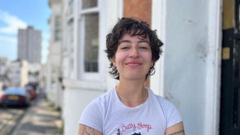 Lola standing in a street, she has curly brown hair and a white t shirt and tattoos