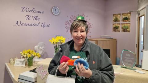 A woman wearing a green hat and grey jumper is holding a number of small knitted hats. She is standing by a desk in a room with purple walls.