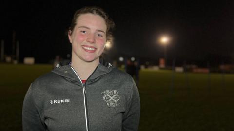 Seren is wearing a grey “Jersey Reds” sports hoodie stands on a rugby pitch at night, smiling at the camera. Floodlights illuminate the field behind her, while the background remains mostly dark.
