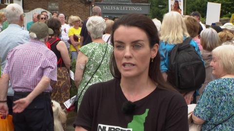 Lucy Williams, who has straight brown hair and wears a black t-shirt, speaks to the camera, while a large group of campaigners gather in the backdrop