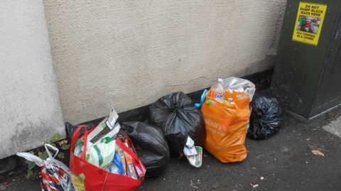 Several tied bags of rubbish are pictured on the floor in a street. A poster warning about fly-tipping is stuck on a green box