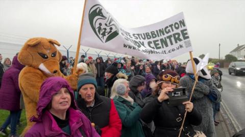 A large throng of protesters by the side of the road. A banner reads: 'Save Stubbington Study Centre'. One protester is dressed as a fox. Most are in winter jackets and hoods.