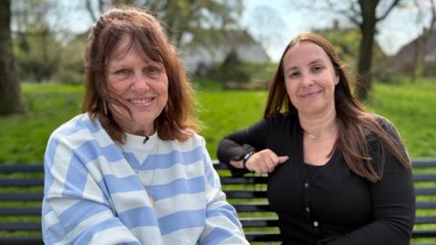 Margaret and Leanne sit on a bench in a park. Both are smiling at the camera. Margaret has short dark hair, and wears a white and purple striped jumper with a gold locket necklace. Leanne has longer dark hair, and is wearing a black cardigan, a black watch and a short silver necklace. 