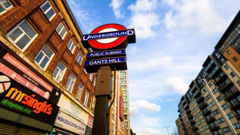 A street-level view of the London Underground sign for Gants Hill station, featuring the red and blue roundel against a bright blue sky with scattered clouds. Surrounding the sign are multi‑storey brick and modern buildings, including shops with colourful signage along the street.