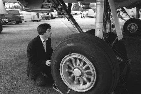 Black and white image from Doctor Who with Patrick Troughton in the foreground hiding behind a plane wheel on a runway