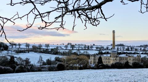 A lovely snowy scene with trees and a large building seen with a dusting of the white stuff. The sky is mostly clear with a few white and grey clouds moving away.