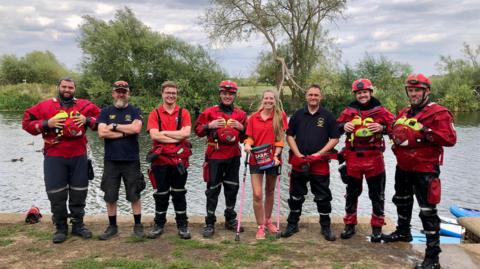 Eight volunteers are standing in front of a river, in various states of dress in their red rescue kits. Three are wearing helmets, many are wearing full dry suits, and all are smiling. 