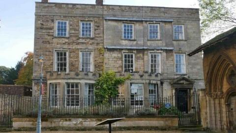 A three-storey pale stone building with panelled window frames and railings outside the property, bordering a path.