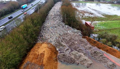 An aerial shot of the dumped waste, stretching out in a road-like line into the distance and surrounded on both sides by trees next to a motorway.