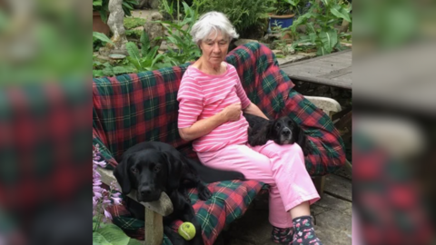 Shirley Pope in a pink t shirt and pink trousers, sitting on a tartan-blanketed bench with two dogs.
