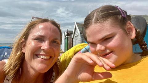 Liz and Ava Chaplin sitting in front of beach huts on a cloudy but warm day. They are both smiling. Liz, who has fair hair, is wearing a white vest top with black stripes, and has sunglasses on her head. Ava is wearing a yellow T-shirt and has her brown hair in plaits.