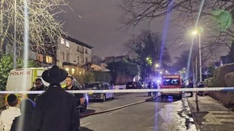 Police tape cordons off scene of fire. Two boys and a man stand on one side with a police van the other side and a red van in the distance 