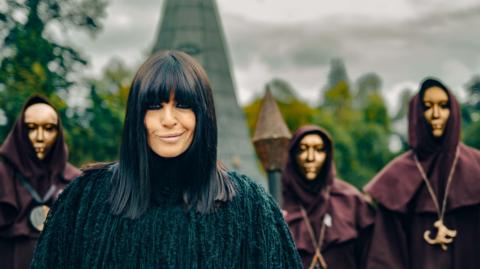 Claudia Winkleman wears a black coat as she smiles at the camera. Behind her is part of a castle and three people in black hoods and golden masks.