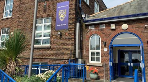 The entrance to Ballakermeen High School, which is a red-and-brown brick building with a blue-framed glass door and blue railings leading up to it.