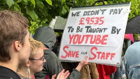 Students standing in the rain at a protest. One is holding up a sign saying: 'We didn't pay £9,535 to be taught by YouTube. Save our staff."