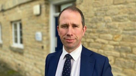 Matthew Barber, a man with short hair and wearing a blue suit, looking straight at the camera with a stone wall behind him.