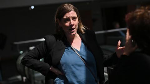 Jess Phillips standing up, wearing blue top underneath black blazer. She has one hand on her hip and is facing someone and talking to them.