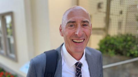Deputy Steve Williams smiles into the camera. He is wearing a grey blazer and a white shirt with a navy tie.