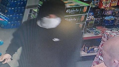 CCTV image inside a shop featuring a hooded man wearing a mask over half his face