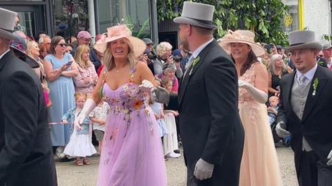 Four couples are dancing in the street, wearing mornign coats and smart frocks . The pavements are lined with spectators.