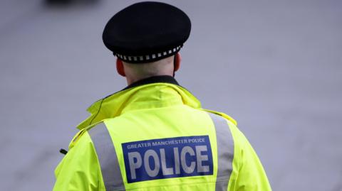 A police officer stands with his back to the camera wearing a police hat, and a hi-vis jacket, with a blue rectangle sign on the back saying 'Greater Manchester Police'