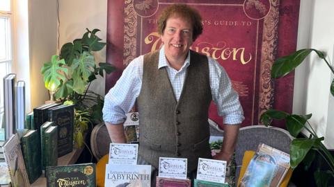 John Tarrow surrounded by a large collection of his books. He is wearing a brown waistcoat over a white shirt.