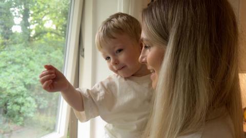 A young woman stands looking out the window, holding a blonde haired boy.