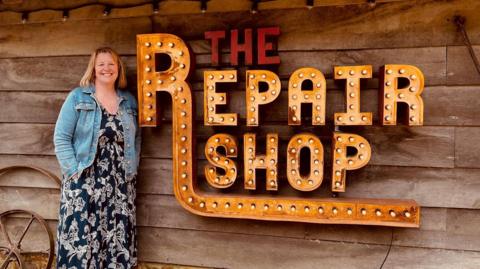 A woman wearing a blue patterened dress and a jean jacket is standing next to a sign saying The Repair Shop. The sign is made of metal and has light bulbs inside. It is hanging on a wooden shed.