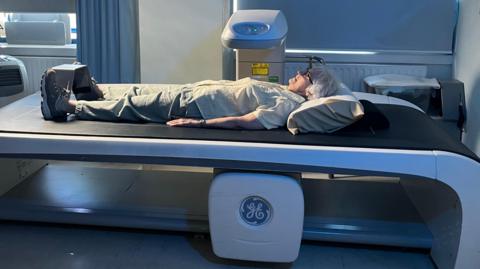 A patient lies flat on a DEXA scanning table in a clinical room, with legs supported and positioned for imaging. The scanning machine sits behind the patient, while curtains, a window, and medical equipment form the background of the softly lit examination space.